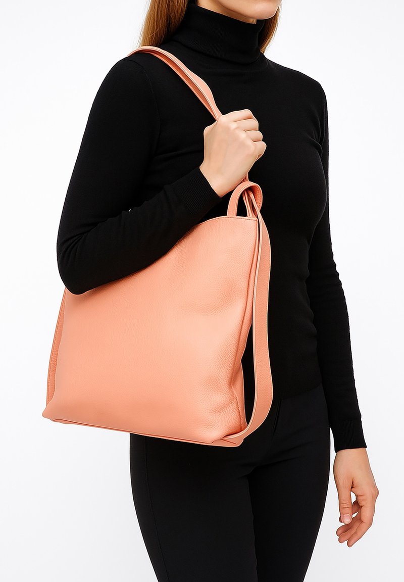 Perzikkleurige leren handtas met een zachte textuur, een afgeronde bovenkant en lange hengsels, vastgehouden tegen een zwarte top en donkere broeken. Eenvoudig en functioneel ontwerp.