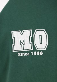 Zöld pamut ing nagy fehér hímzéssel, amelyen a "MO" és alatta a "Since 1998" felirat olvasható. Egyszerű, sportos dizájn kontrasztos színekkel.