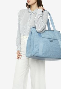 Sac fourre-tout en toile bleue avec un tissu texturé et des anses lisses, présentant une poche avant et un design structuré, tenu par une personne portant une chemise rayée.
