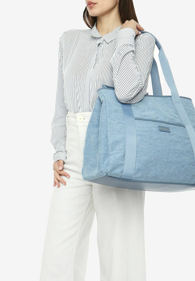 Sac fourre-tout en toile bleue avec un tissu texturé et des anses lisses, présentant une poche avant et un design structuré, tenu par une personne portant une chemise rayée.
