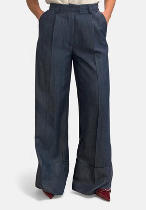 Pantaloni grigio scuro a gamba larga con pieghe frontali, indossati con décolleté bordeaux a punta e mani nelle tasche.