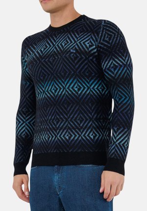 Uomo che indossa un maglione nero e blu con motivi geometrici, maniche lunghe e jeans blu, in piedi davanti a uno sfondo semplice.