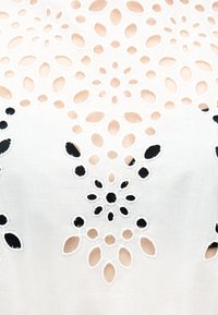 Λευκό ύφασμα με συμμετρικά κεντήματα λουλουδιών και κοψίματα που αποκαλύπτουν μπεζ και μαύρα φόντα μέσα από τις τρύπες.