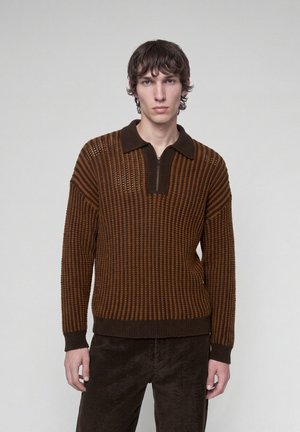 Giovane uomo con capelli castani corti che indossa un maglione di maglia marrone e arancione con colletto e cerniera, in posa davanti a uno sfondo chiaro e semplice.