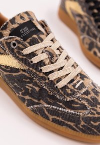 Sneaker sportivo con tessuto in stampa leopardata beige e nera, lacci beige, striscia dorata e suola in gomma marrone chiaro su uno sfondo bianco.