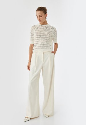 Weißer Pullover aus Strick mit kurzen Ärmeln, strukturiertem Muster, kombiniert mit cremefarbenen, weit geschnittenen Hosen und weißen spitzen Ballerinas. Minimalistisches Design.