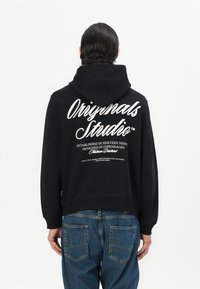 Black hooded sweatshirt with a cropped design. White text on the back includes "Originals Studio," established details, and a tagline.