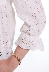 Witte eyelet blouse met een geschulpte onderkant en puffed mouwen. Een gouden, golvende armband siert de pols.