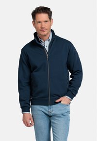 Marineblauwe ritsjack met staande kraag, elastische manchetten en zijzakken. Gecombineerd met een gestreept shirt, gedragen met lichtblauwe jeans.