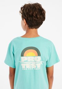 Tyrkysové bavlněné tričko s retro duhovou grafikou v oranžové, žluté a černé barvě s textem "PRO TEST SUN SEA SURF" níže.