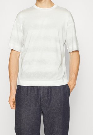 Weißes T-Shirt mit kurzen Ärmeln und Rundhalsausschnitt, versehen mit dezenten Streifen. Glatte Textur, legerer Schnitt, kombiniert mit dunkler, leicht strukturierter Hose.