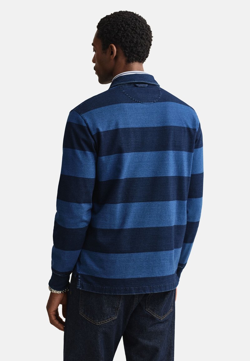 Uomo di spalle che indossa una maglia da rugby a maniche lunghe con ampie strisce orizzontali blu navy e blu e jeans in denim scuro.