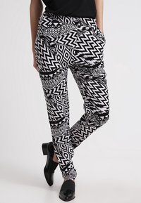 Black and white patterned pants feature geometric designs, a relaxed fit, side pockets, and a cuffed hem. They are worn with black shoes.