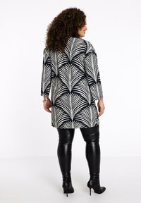 Zwart-wit gestreepte lange jas met een loofontwerp, gecombineerd met glanzende zwarte leggings en hoge zwarte schoenen.