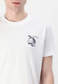 Joven llevando una camiseta blanca con un pequeño gráfico de un limón negro y el texto "JOOP! JEANS" en el lado izquierdo del pecho.