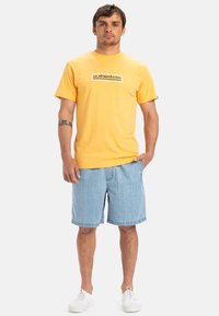 Geel shirt met korte mouwen met een grafische print op de voorkant, gecombineerd met denim shorts en witte sneakers. Eenvoudig ontwerp en casual pasvorm.