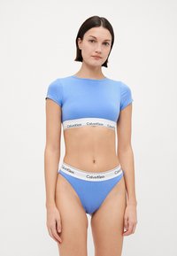 Mėlynas medvilninis trumpas viršus su trumpomis rankovėmis, derinamas su atitinkamais mėlynais stringais. Yra baltas juosmuo su "Calvin Klein" logotipu.