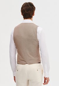 Kamizelka w brązowy wzór houndstooth o prostym kroju, nałożona na białą koszulę, uzupełniona jasnymi spodniami. Brak widocznych zapięć.