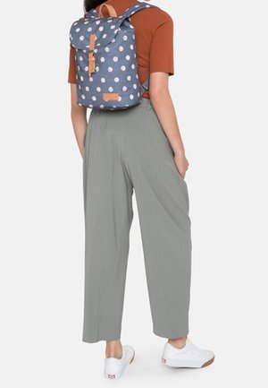 Batoh tmavě modré barvy s růžovými puntíky, doplněný opálenými koženými detaily a nastavitelnými popruhy. Modelka má na sobě šedé kalhoty s širokými nohavicemi a bílé tenisky.