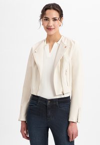 Crème leren cropped jack met zilveren rits, drukknoopjes en gestructureerde schouders, gecombineerd met een witte top en donkerblauwe jeans.
