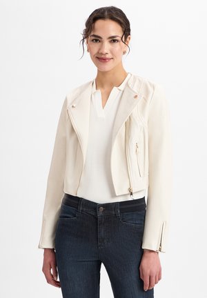 Crème leren cropped jack met zilveren rits, drukknoopjes en gestructureerde schouders, gecombineerd met een witte top en donkerblauwe jeans.