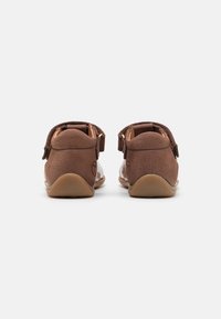 Brązowe zamszowe buty o gładkim wykończeniu, z regulowanymi paskami i zaokrąglonym stylem noska. Lekkie gumowe podeszwy widoczne z tyłu.