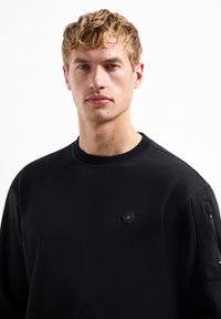 Zwarte trui met een ronde hals, voorzien van een klein zeszijdig logo en een met rits afgesloten zak aan de linkermouw. Zachte, getextureerde stof.