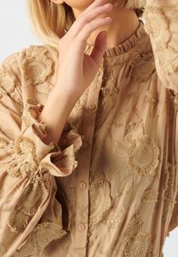 Sheer beige blouse with floral embroidery, featuring ruffled cuffs and a high neck. Fastened with buttons, showcasing textured fabric details.