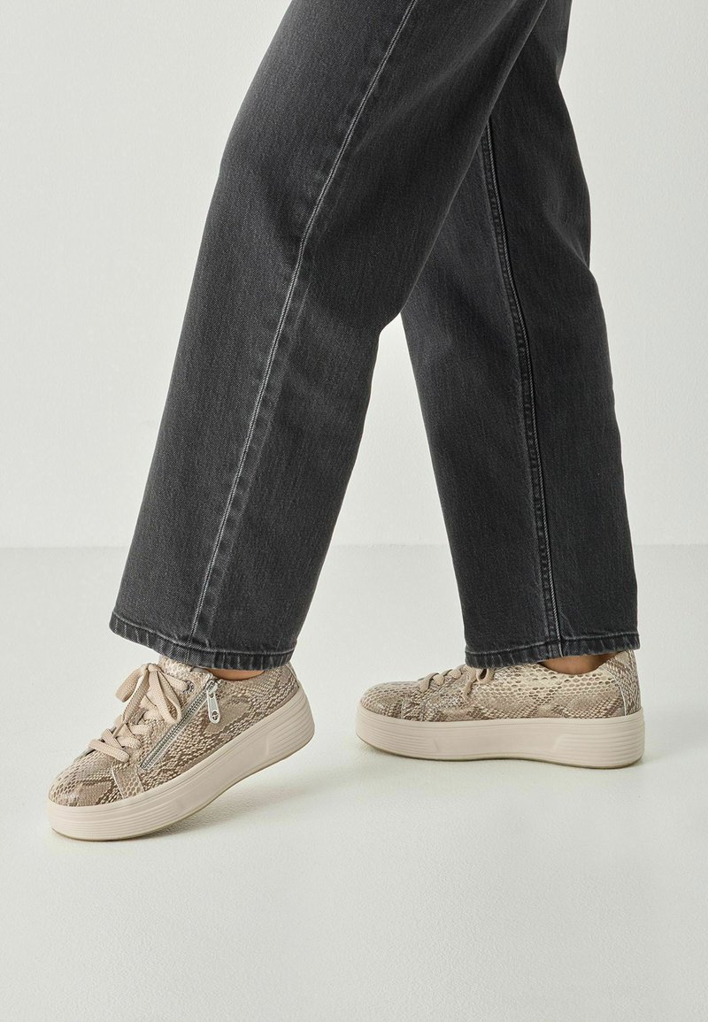 Béžové tenisky s texturovaným hadím vzorem, šněrováním a masivní bílou podešví. Nošeny s černými džínami.