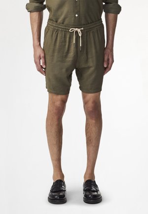 Mężczyzna noszący oliwkowozielone szorty ze sznurkiem, dopasowaną podwiniętą koszulę i czarne skórzane mokasyny stojący na białym tle.