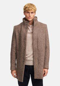 Abrigo de mezcla de lana marrón con cuello alto, tejido texturizado, cierres de botones y bolsillos laterales, llevado sobre un suéter beige.