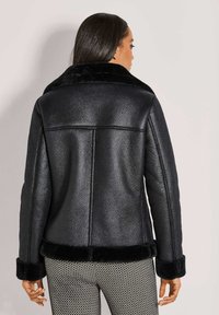 Zwarte leren jas met een zachte zwarte bontkraag, gestructureerde afwerking en gedetailleerde stiksels. Heeft brede manchetten en een kort model.