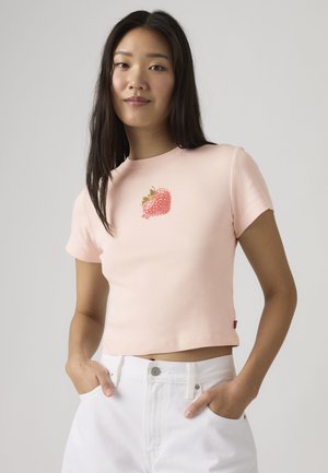 Mlada ženska nosi svetlo roza kračko majico s sliko jagode in bele visoko pasne hlače, roke v žepih.