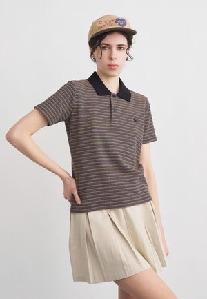 Νέα γυναίκα που φοράει ριγέ πόλο πουκάμισο με μαύρο γιακά, μπεζ πλισέ φούστα και καφέ καπέλο μπέιζμπολ με κείμενο, στέκεται με το χέρι στη μέση.