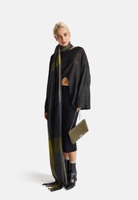 Wielki sweter w kolorze węgla z bocznym wycięciem, zestawiony z ciemno wzorzystym szalikiem z zielonymi akcentami, czarną spódnicą i botkami za kostkę.