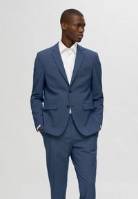 Costume ajusté bleu en tissu texturé, avec une veste à un bouton dotée de deux poches avant et un pantalon assorti. Chemise blanche en dessous.