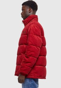 Rød kordfløjl puffjakke med h�øj krave, quiltet design og broderet logo på brystet. Har lynlåslukning og sidelommer.