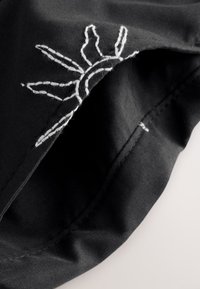 Tessuto nero con una texture morbida, decorato con un ricamo bianco raffigurante un sole vicino al bordo, che mette in risalto le cuciture dettagliate.