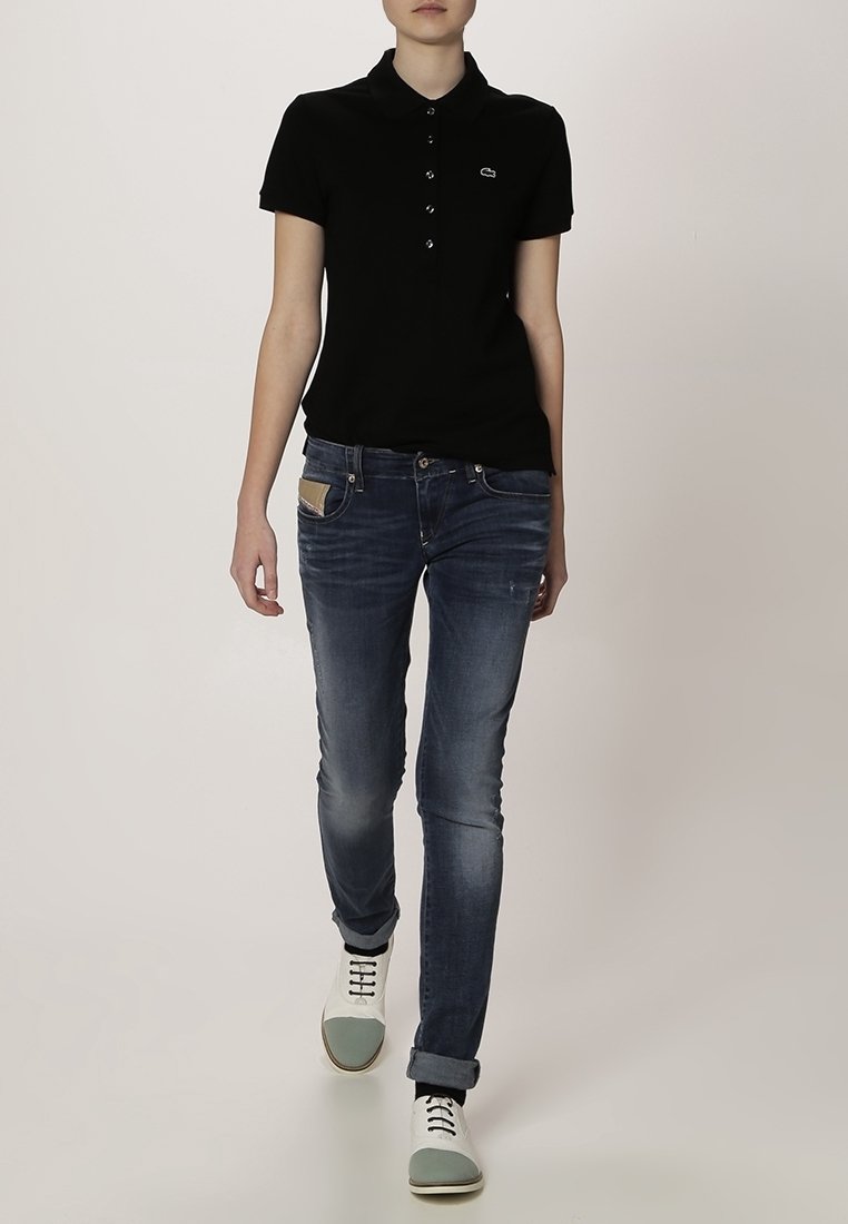 Personne marchant portant un polo noir à manches courtes, un jean bleu délavé avec des revers, et des chaussures blanches avec des embouts gris clair.