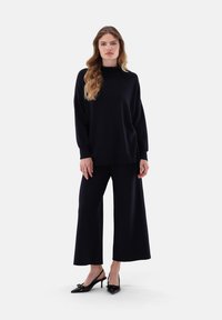 Marineblaues, übergroßes Sweatshirt mit hohem Kragen, weiten Hosen und schwarzen spitzen Absätzen; glatte Textur, lockere Passform und minimalistisches Design.