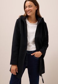 Veste polaire noire avec fermeture à glissière sur le devant, capuche et cordons de serrage. Dotée d'un tissu texturé et de poches latérales, elle est associée à un t-shirt blanc et à un jean.