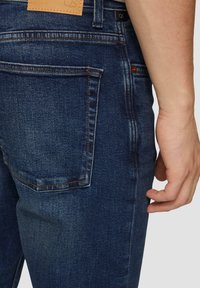 Jeans de mezclilla azul oscuro con corte slim, que cuentan con costuras naranjas, una etiqueta de cuero en la cinturilla y detalles visibles en los bolsillos traseros.
