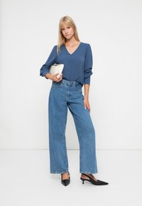 Blå langermet topp med V-hals, parret med vide blå jeans. Hun holder en kremfarget clutch og har på seg svarte hælesandaler.