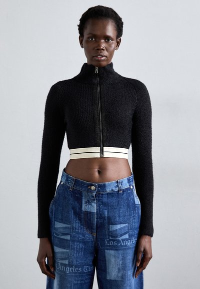 Vrouw in een zwarte cropped zip-up trui met witte details en hoge taille blauwe jeans met krantenprint tekst.