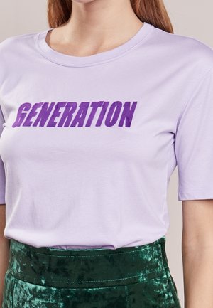 Frau trägt ein hellviolettes T-Shirt mit kurzen Ärmeln, auf dem "GENERATION" in fetten lila Buchstaben gedruckt ist, und grüne Samthose.