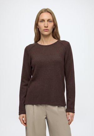 Vrouw met steil blond haar draagt een donkerbruin shirt met lange mouwen en beige broek, staand tegen een effen lichte achtergrond.