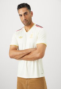 Maglietta da calcio color crema chiaro con scollo a V, maniche corte, dettagli a strisce nere e rosse e particolari dorati, realizzata in un tessuto morbido.
