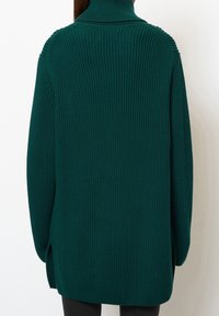 Grüner, oversized gestrickter Pullover mit hohem Kragen, der eine gerippte Textur und seitliche Schlitze aufweist. Keine sichtbaren Verschlüsse oder Verzierungen.