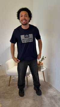 Mann mit kurzen lockigen Haaren trägt ein schwarzes "Weekend Offender" T-Shirt, schwarze Hose und schwarze Schuhe und steht neben einem weißen Stuhl und einem Beistelltisch.