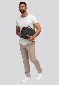 Bolso tipo mensajero sintético negro con cierre de cremallera, asa superior y detalle del logo, sostenido por una persona que lleva una camisa blanca ajustada y pantalones claros.